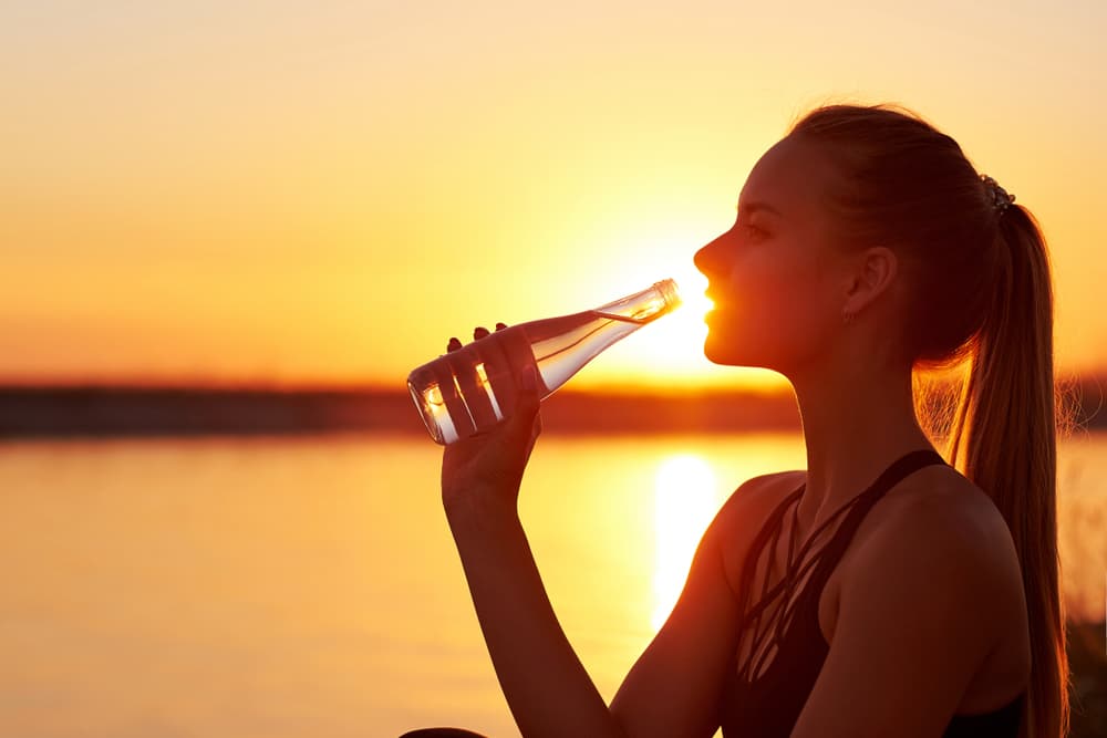 woman drinking water on a beach with the sunset in the background